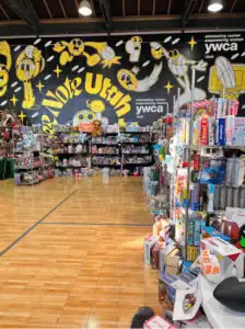 A welcoming toy store with shelves of toys and games, featuring a "Vote Utah" and "YWCA" mural, reflecting community care.