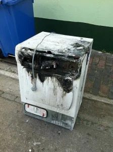 A damaged washing machine sits on a sidewalk by a blue trash bin, illustrating the need for expert, reliable home repair services.