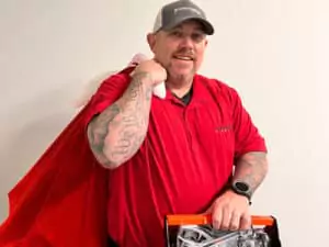 Friendly technician in a red shirt and gray cap holds a cape and toolbox, ready to provide 24/7 expert home repair services.