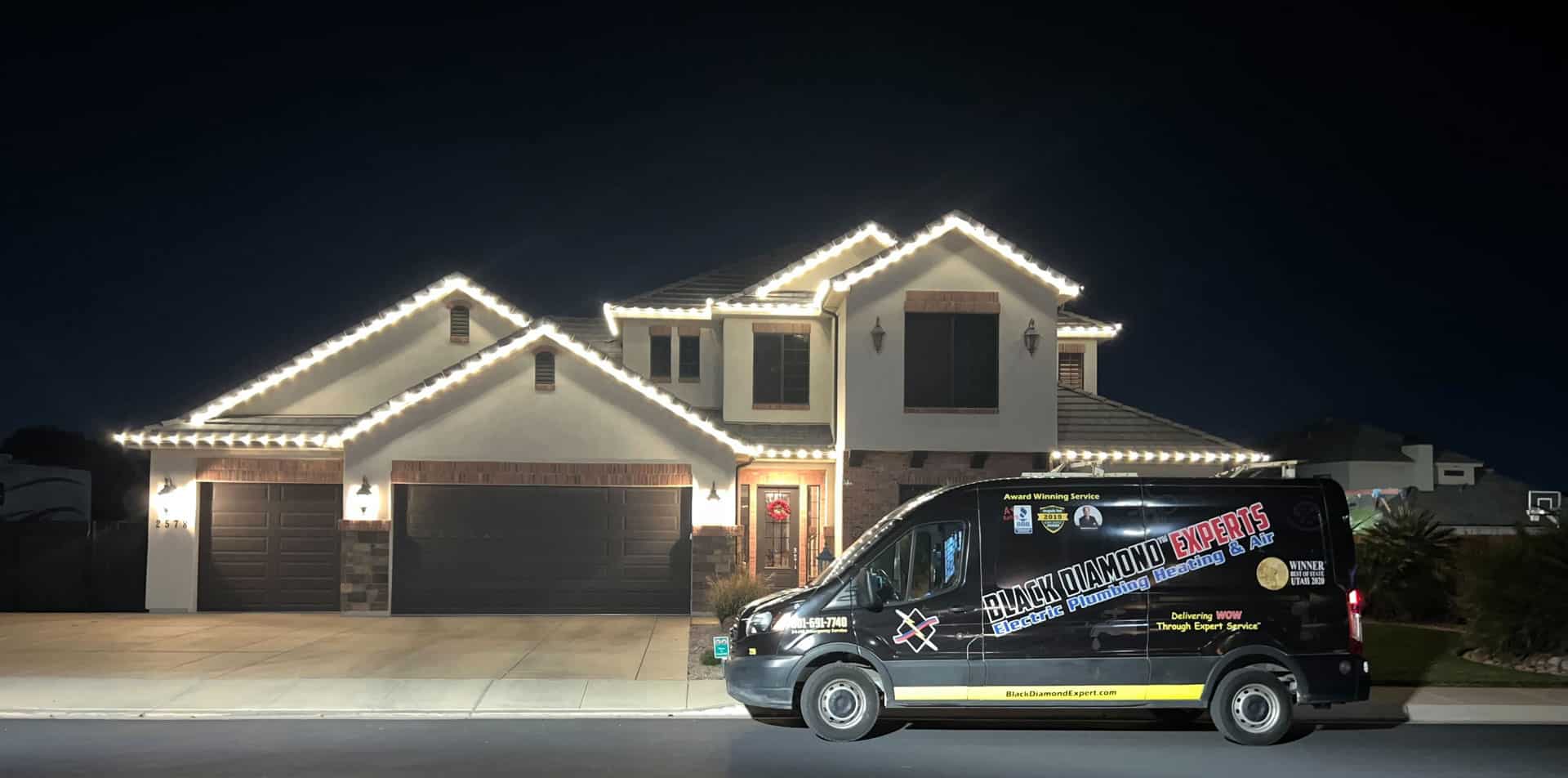 A Black Diamond Experts van sits outside a warmly lit Utah home at night, ready for 24/7 expert plumbing, HVAC, and electrical help.