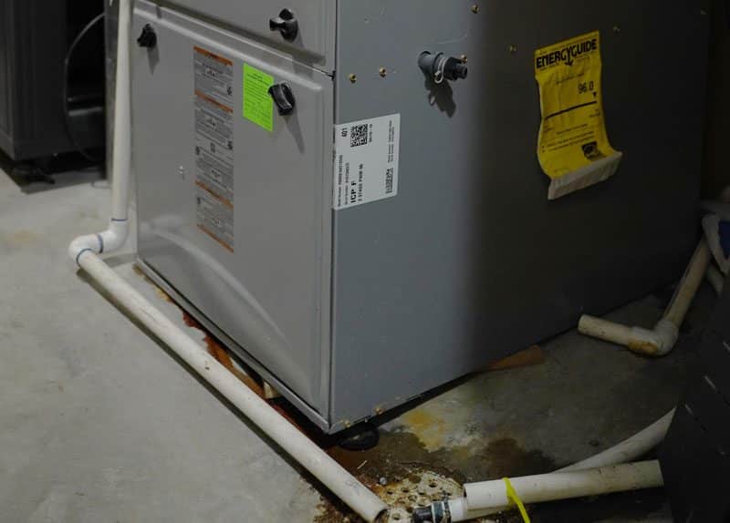 A furnace stands on a concrete floor with white PVC pipes; a rust-colored water stain nearby signals a need for expert, reliable care.