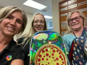 Three women, smiling warmly, show off colorful backpacks—one with a playful pizza design—reflecting trusted, reliable service.