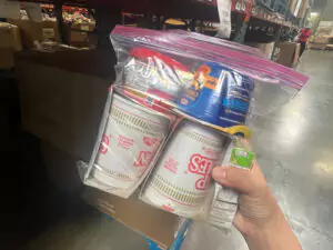 A caring hand holds a clear bag with instant noodles, canned food, and snacks—symbols of reliable home comfort and safety.